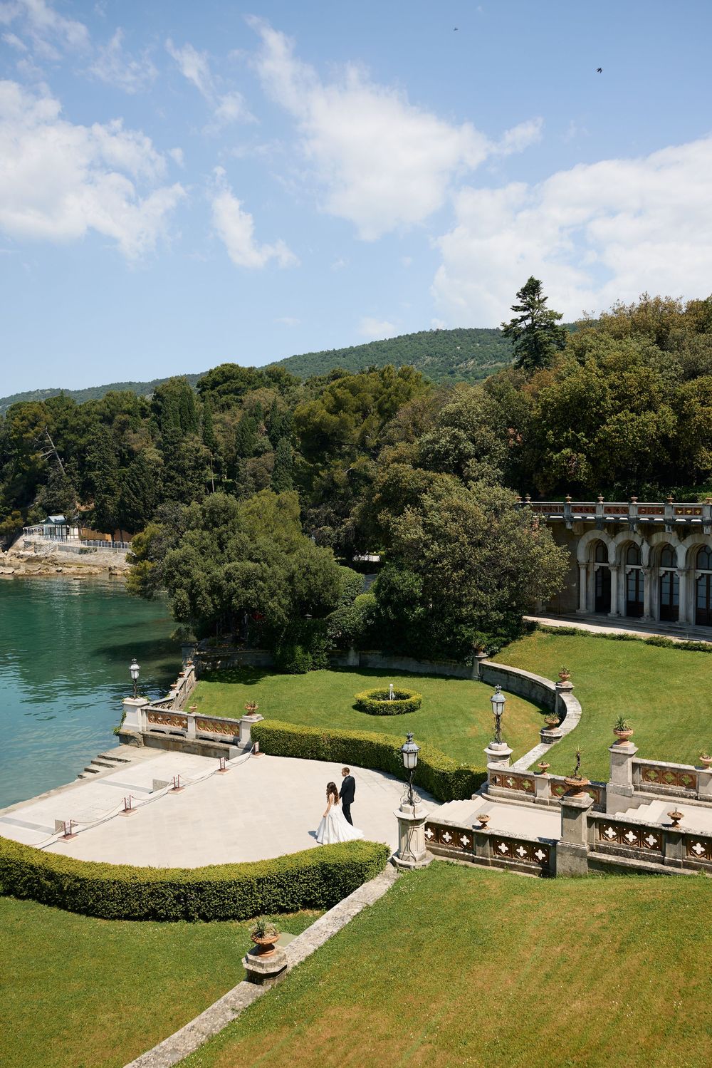 Aerial view of a wedding couple strolling through elegant Italian villa gardens beside turquoise waters.
