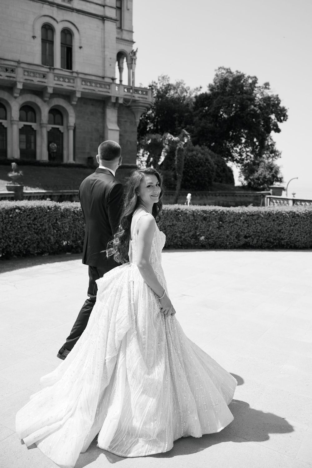 Black and white photo of bride in flowing gown glancing back as groom leads her past grand castle.