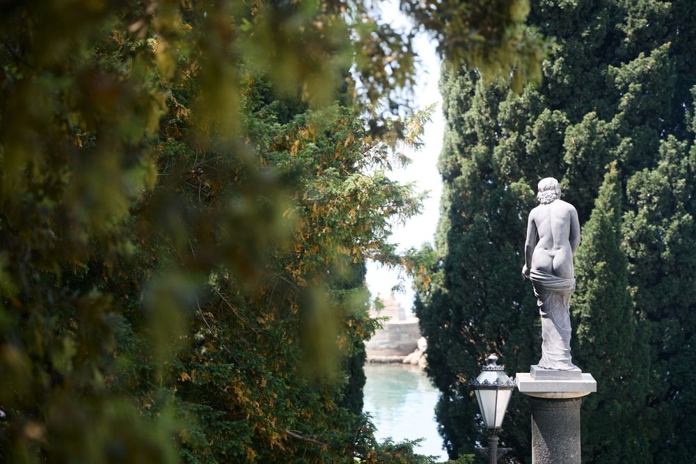 White marble statue surrounded by lush green cypress trees in a serene garden with a decorative lamp post.