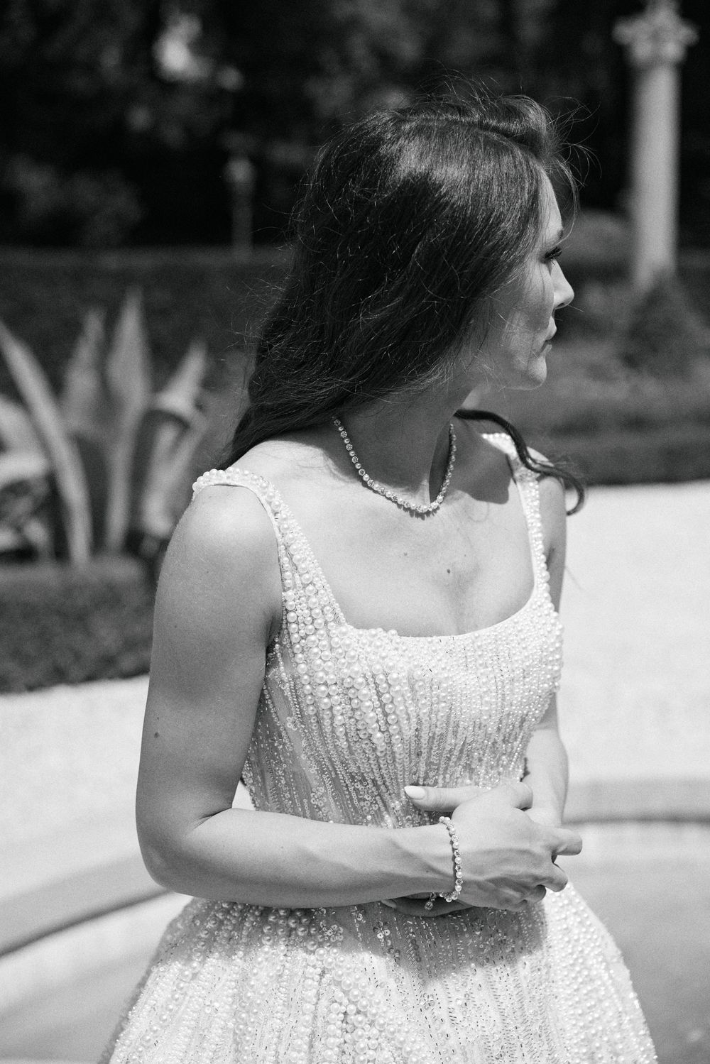 Elegant bride in pearl necklace and beaded white gown poses outdoors in black and white photo.