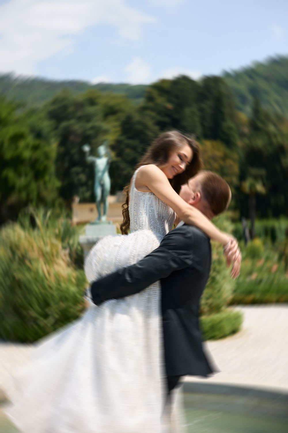 Groom lifting bride in white dress outdoors, surrounded by lush greenery and a statue, captured with a romantic blur effect.