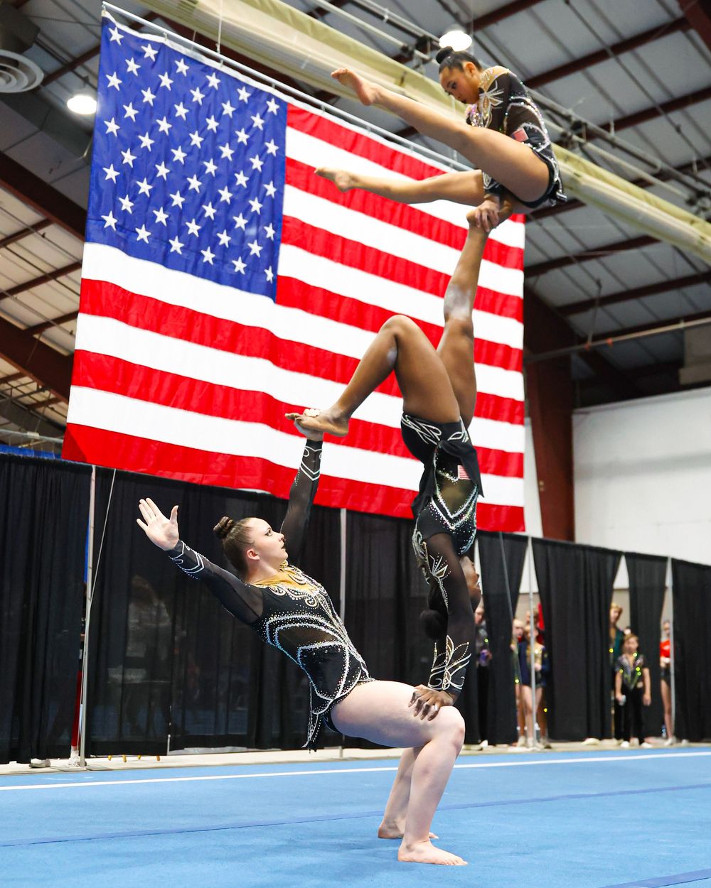 Two gymnasts perform an acrobatic balance routine in front of a large American flag at an indoor gymnastics competition.