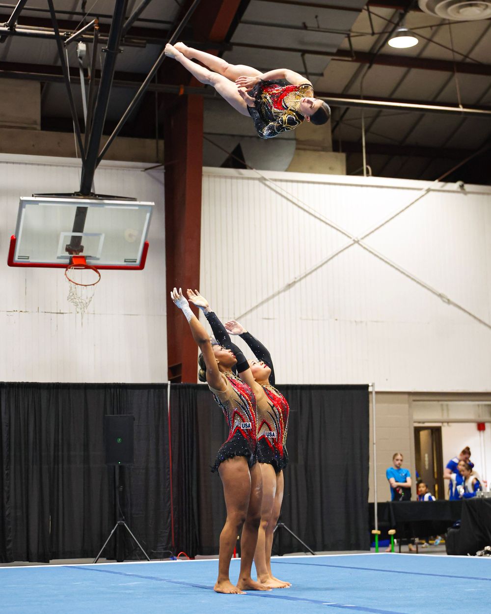Acrobatic gymnasts performing a group balance stunt indoors, with one athlete soaring high above teammates in matching red uniforms.