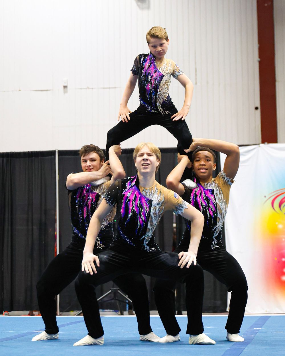 Four acrobatic gymnasts in matching black and purple costumes perform a pyramid formation on a blue mat.