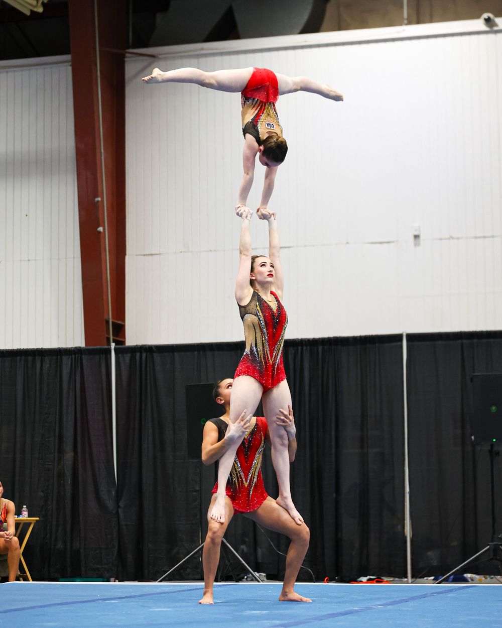 Acrobatic gymnasts perform a stunning three-person balance pyramid in red leotards at an indoor gymnastics competition.