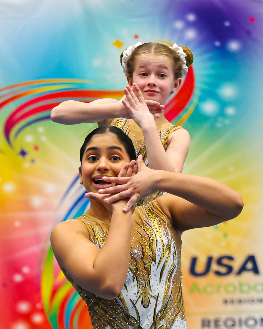 Two young girls perform acrobatic gymnastics at a USA Acrobatics regional competition in sparkly gold costumes.