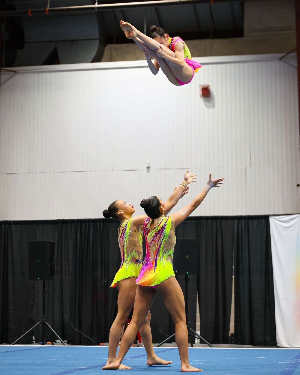 Three acrobatic gymnasts in neon yellow and pink leotards perform a dynamic balance routine, with one athlete soaring high in the air.