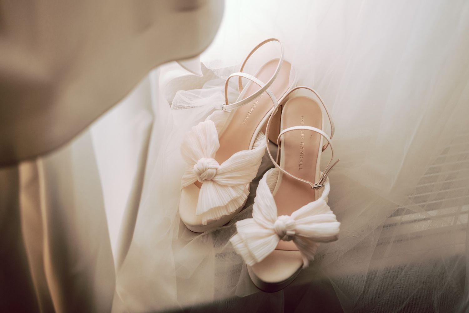 Elegant white bridal sandals with bow details and ankle straps resting on a soft wedding dress.