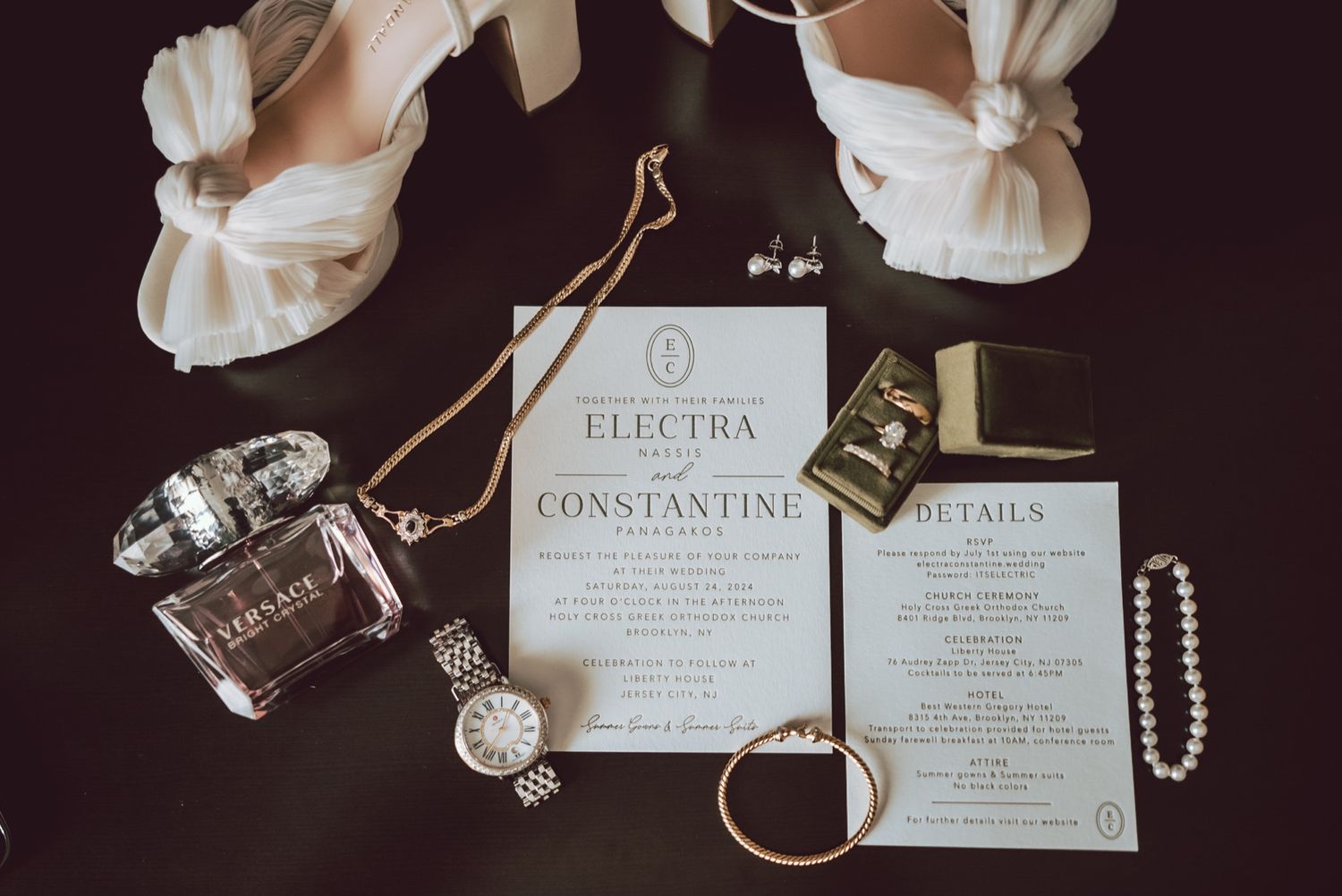 Elegant wedding flat lay with invitation suite, pearl necklace, perfume, watch, gold rings, and bridal heels on dark surface.