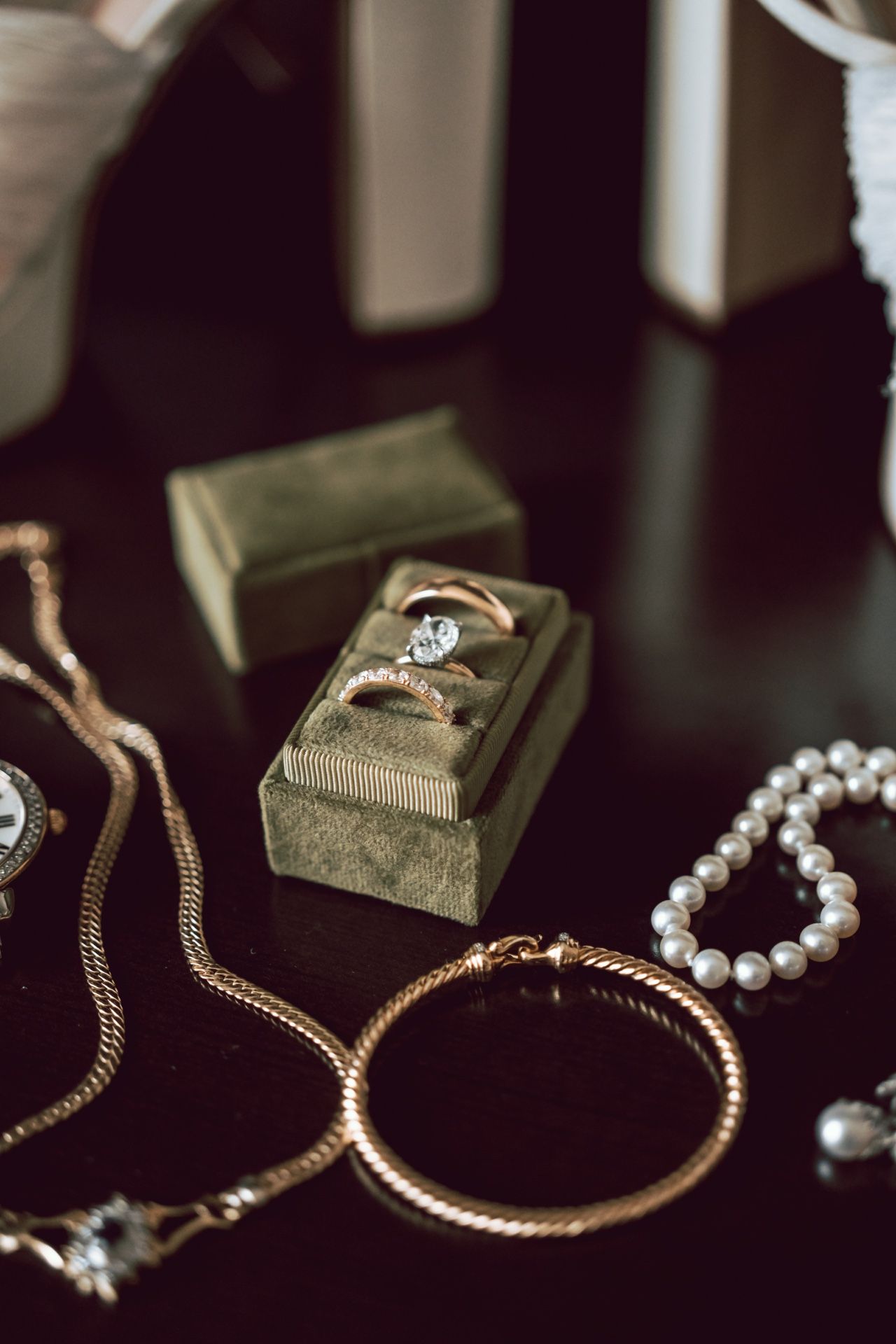 Elegant bridal jewelry flat lay featuring diamond rings in green velvet box, gold bangles, pearl bracelet, and vintage watch.