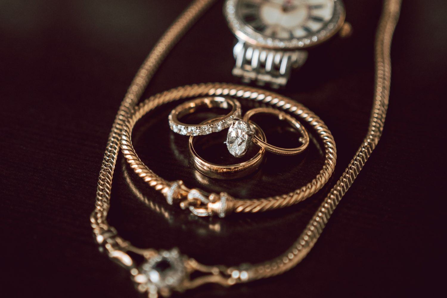 Gold rings and chain necklace arranged elegantly on a dark surface with a luxury watch in the background.
