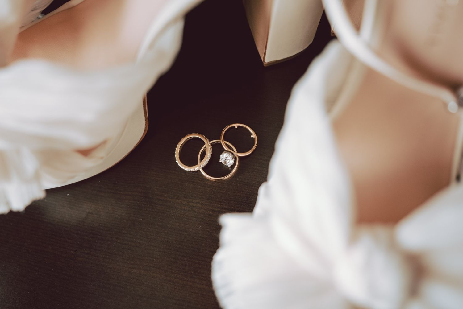 Three gold wedding and engagement rings with diamond displayed between white bridal heels on dark surface.