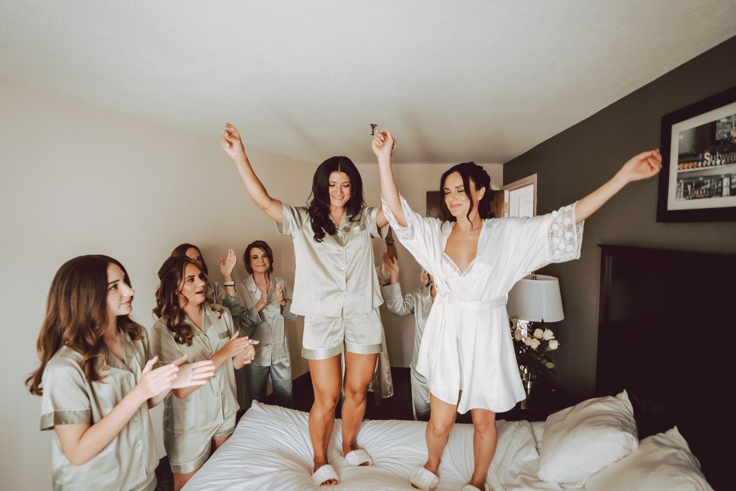 Bridesmaids in matching satin robes celebrate with bride jumping on hotel bed during wedding morning preparations.