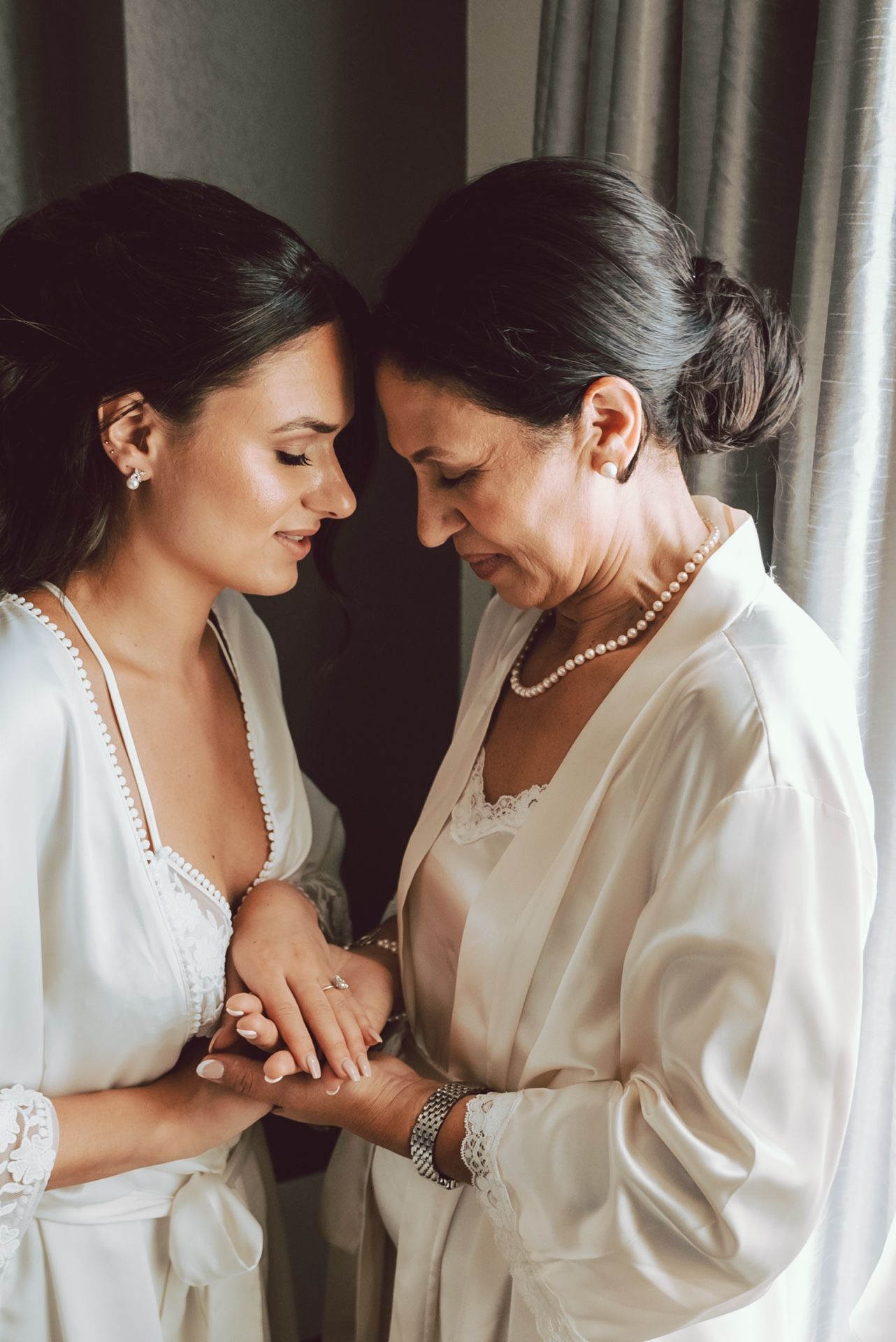 Mother and bride in white robes share an intimate moment holding hands before the wedding ceremony.