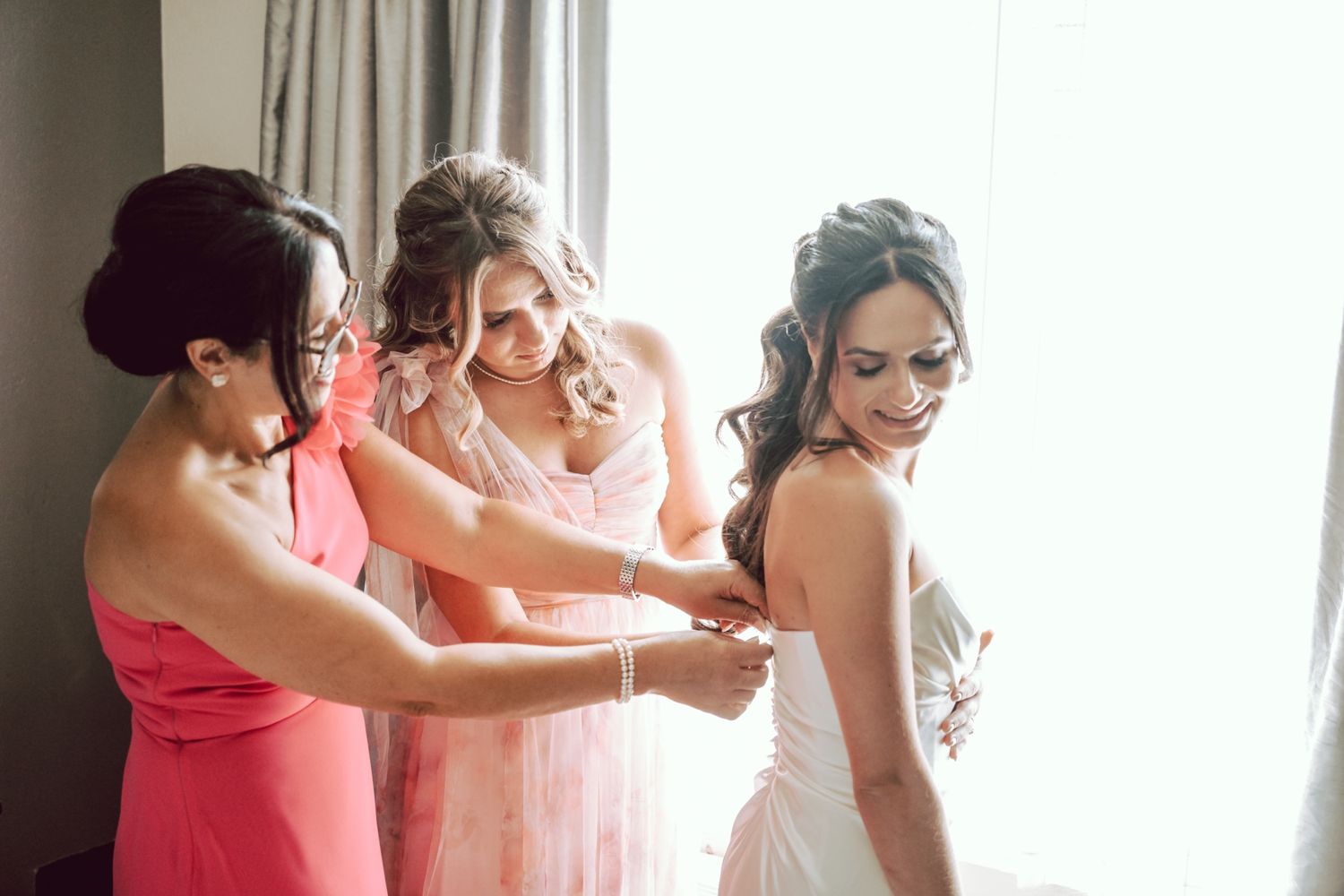 Bridesmaids helping bride get dressed in white wedding gown near window on wedding day.
