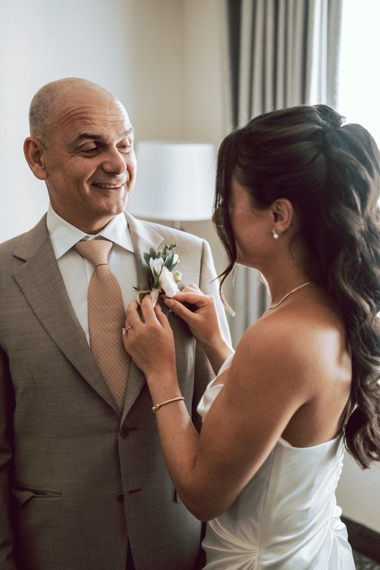 Bride pinning a white boutonniere on a smiling bald groom in a gray suit and pink tie on wedding day.
