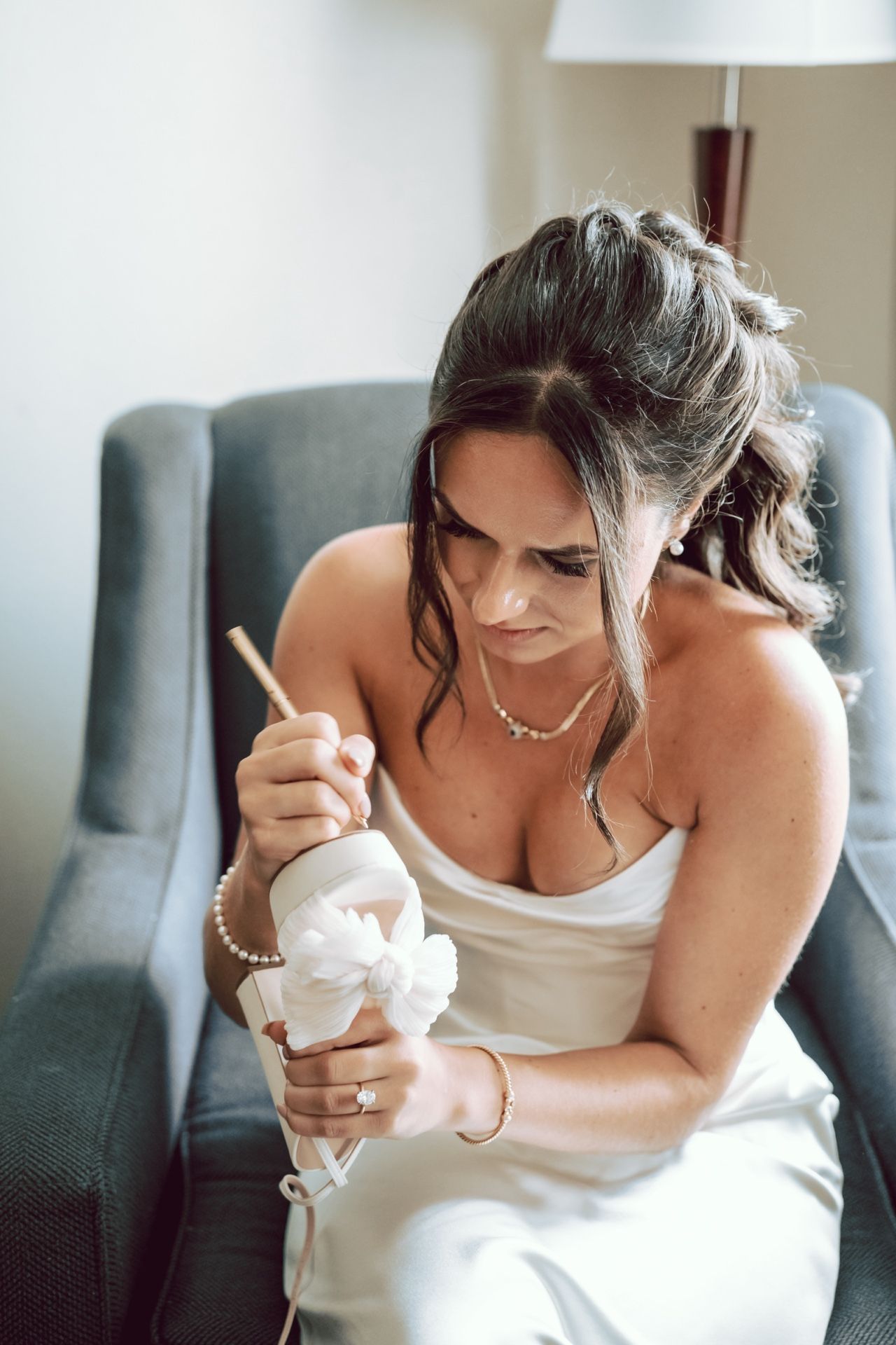 Bride in white gown sitting in gray chair, carefully holding decorated wedding shoe with pearl details.
