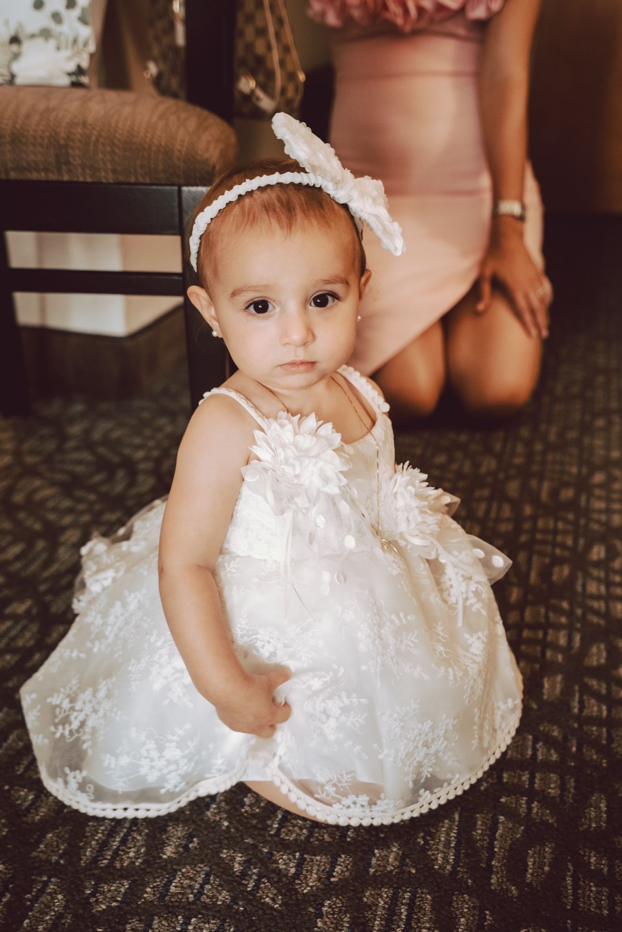 Adorable baby girl in white lace dress and headband sitting on floor during a special occasion.