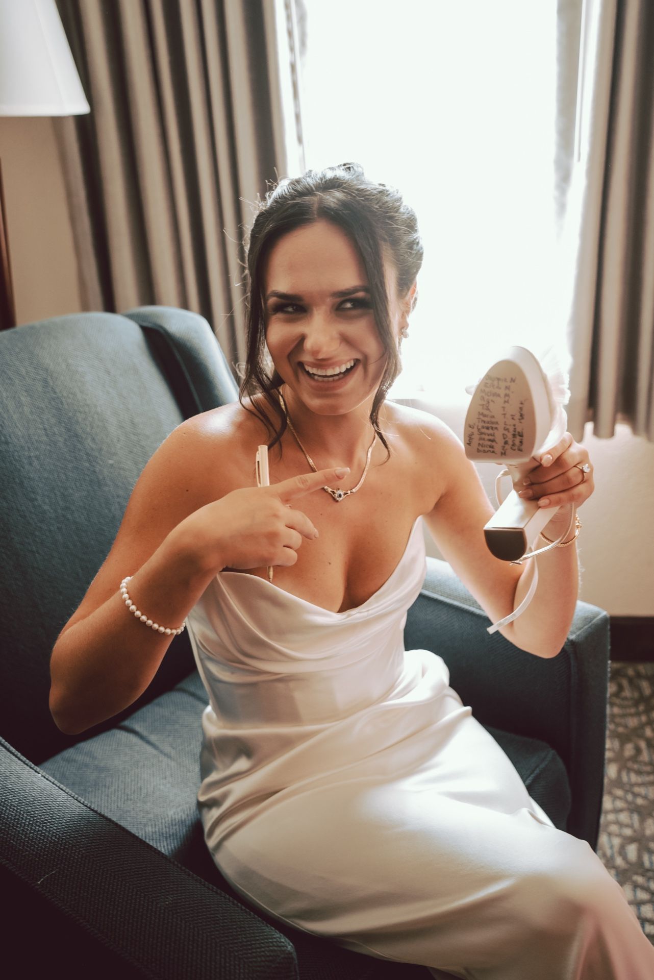 Smiling bride in white satin gown holding a compact mirror while sitting in a blue armchair.