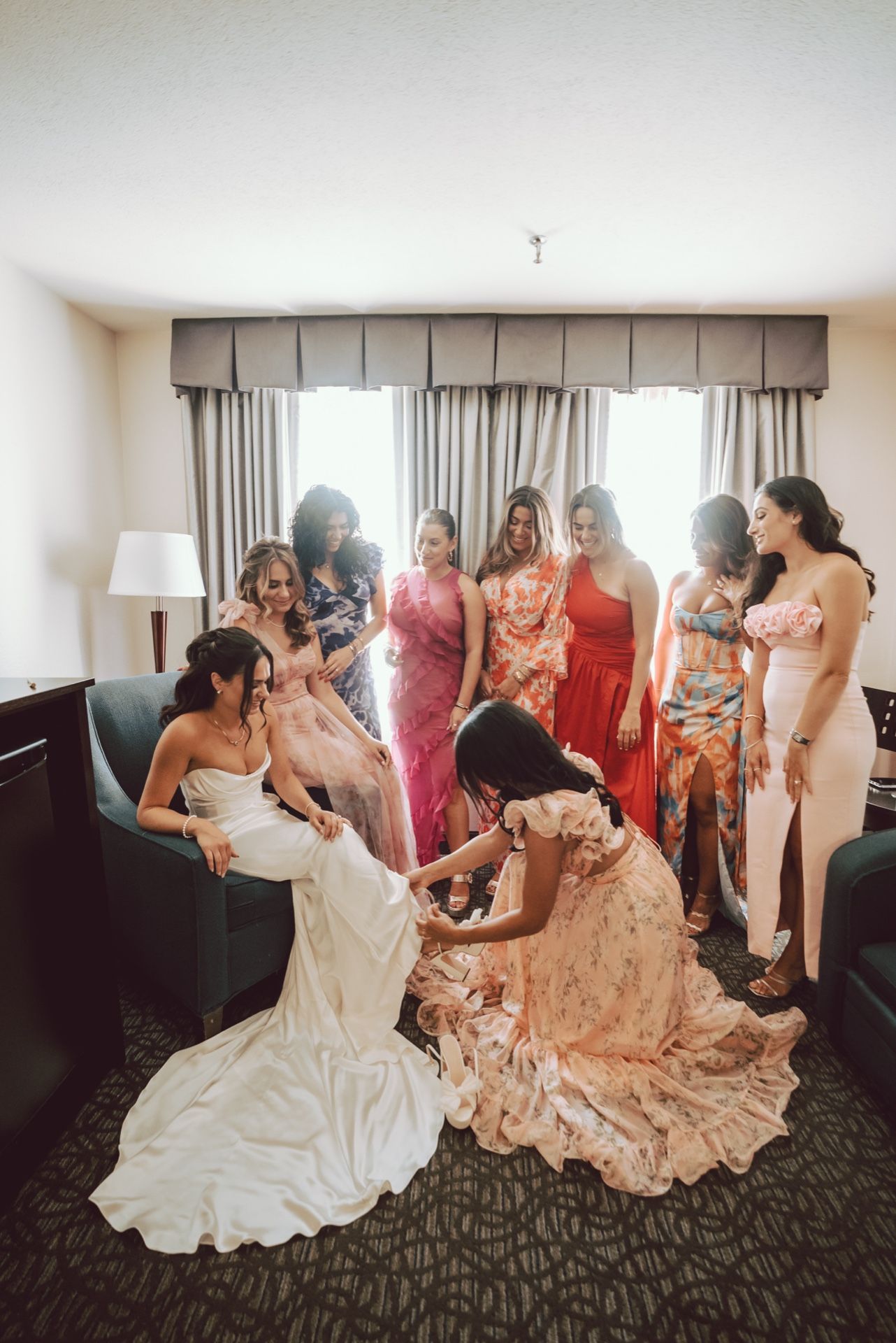 Bride surrounded by bridesmaids in colorful dresses getting shoes put on in hotel room.