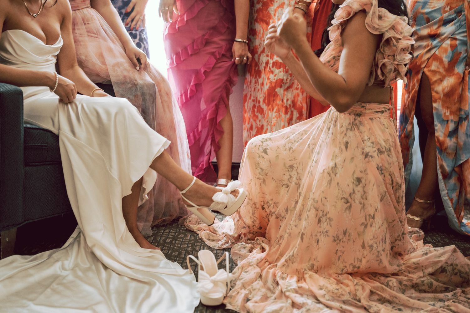 Bridesmaid in floral dress helps bride put on white heels during wedding preparations.