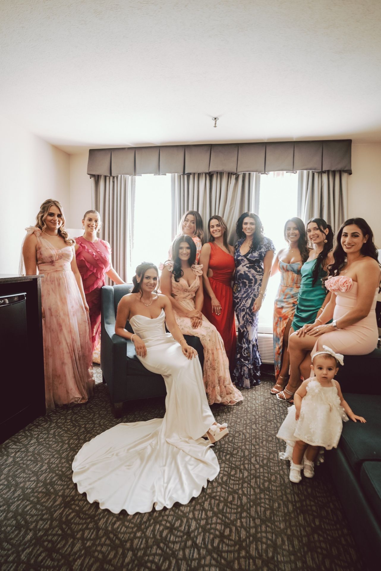 Bride poses with bridesmaids and flower girl in vibrant dresses in hotel room before wedding.