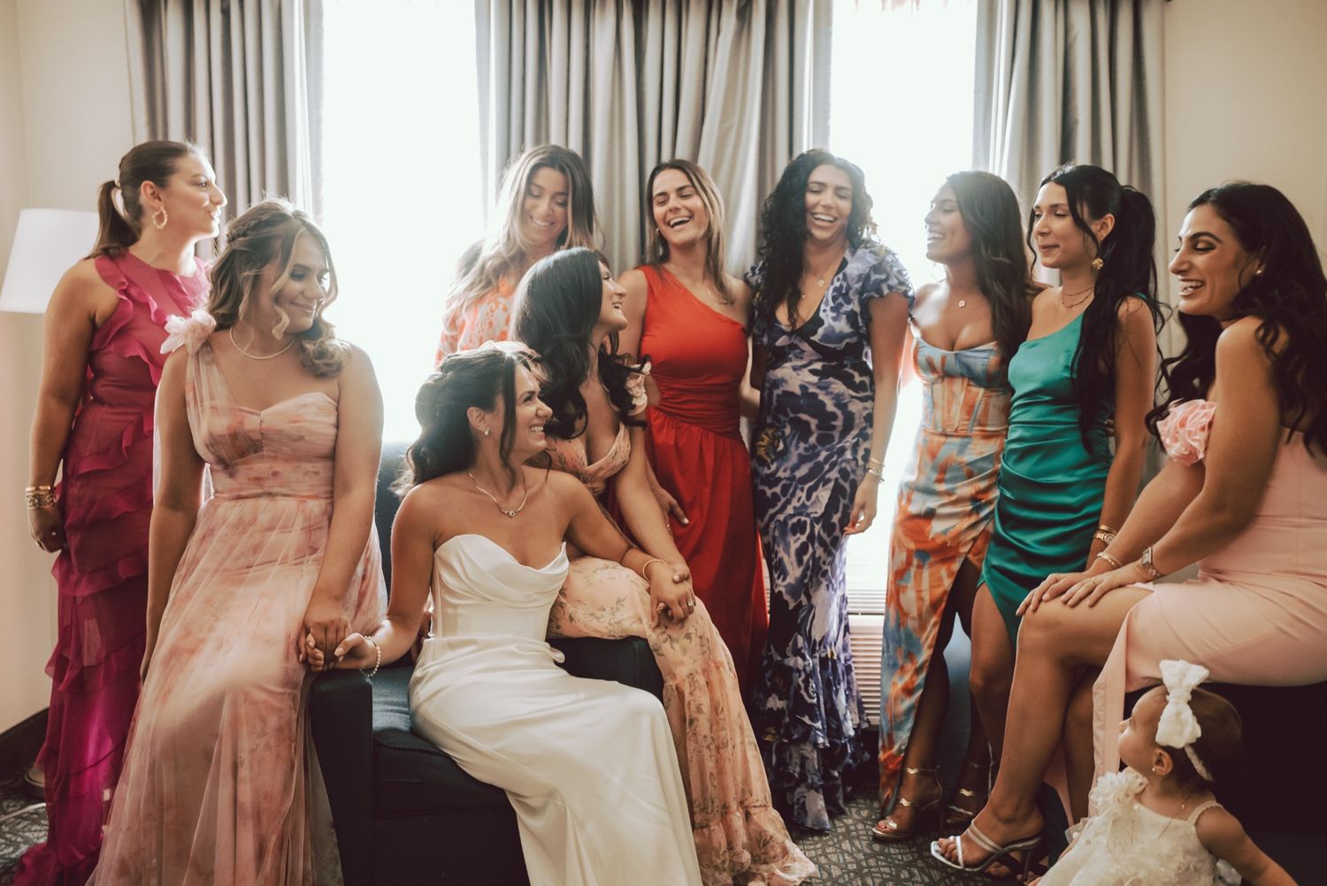 Bride in strapless gown laughs with bridesmaids in colorful dresses during wedding prep.