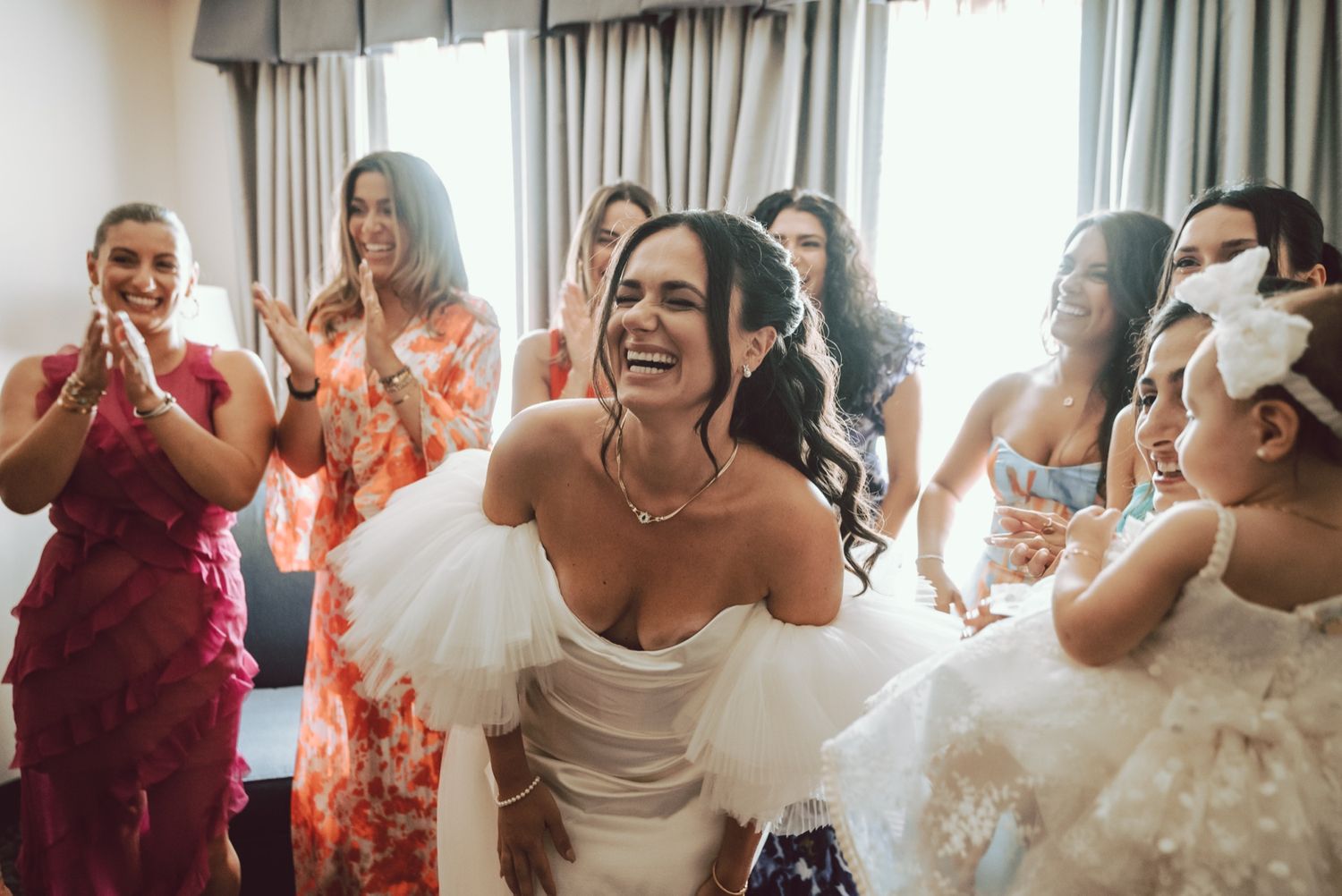 Bride in white dress laughing joyfully with bridesmaids in colorful gowns during wedding preparations.