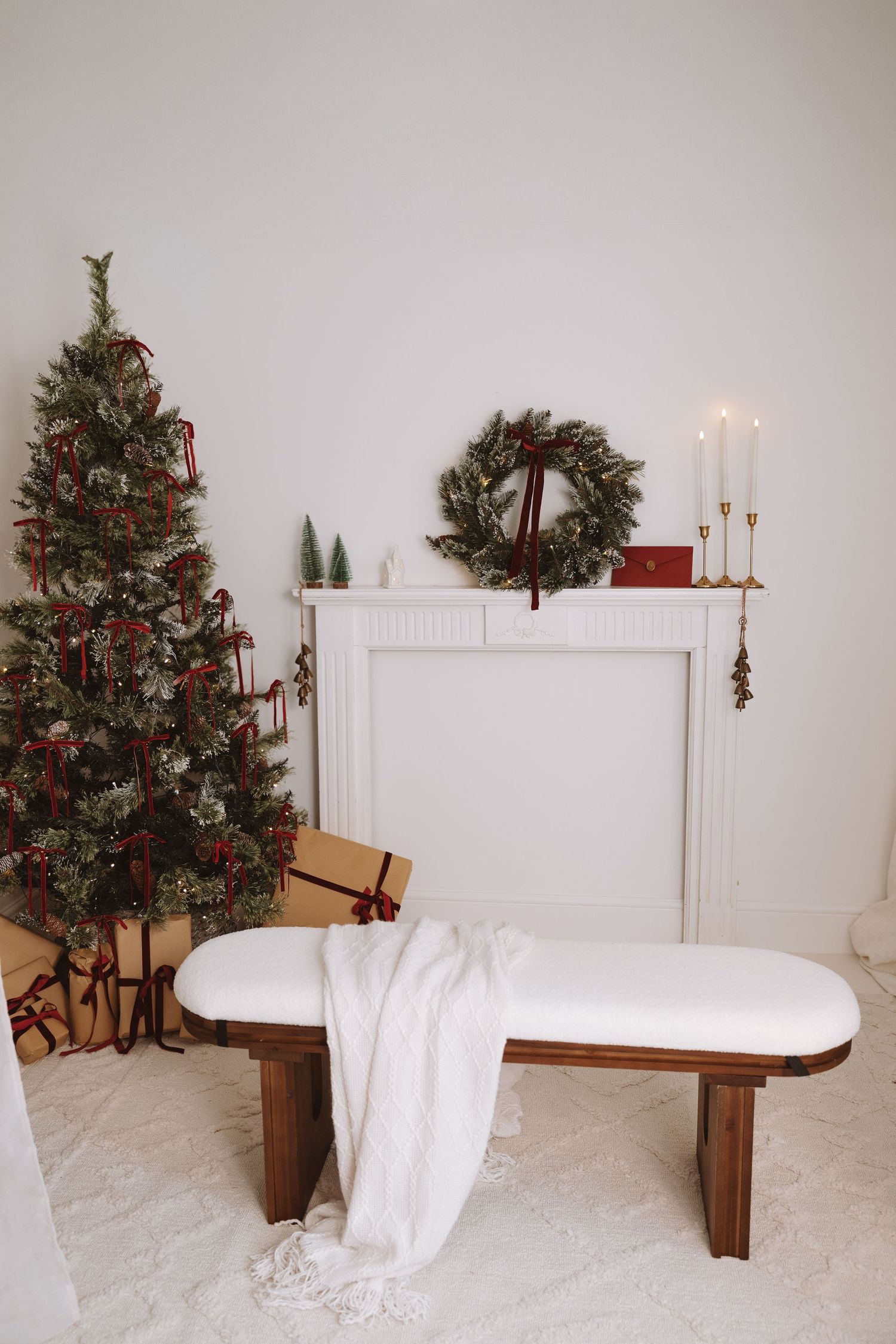 Elegant Christmas room with decorated tree, wrapped gifts, white bench, and wreath above fireplace mantel with candles.