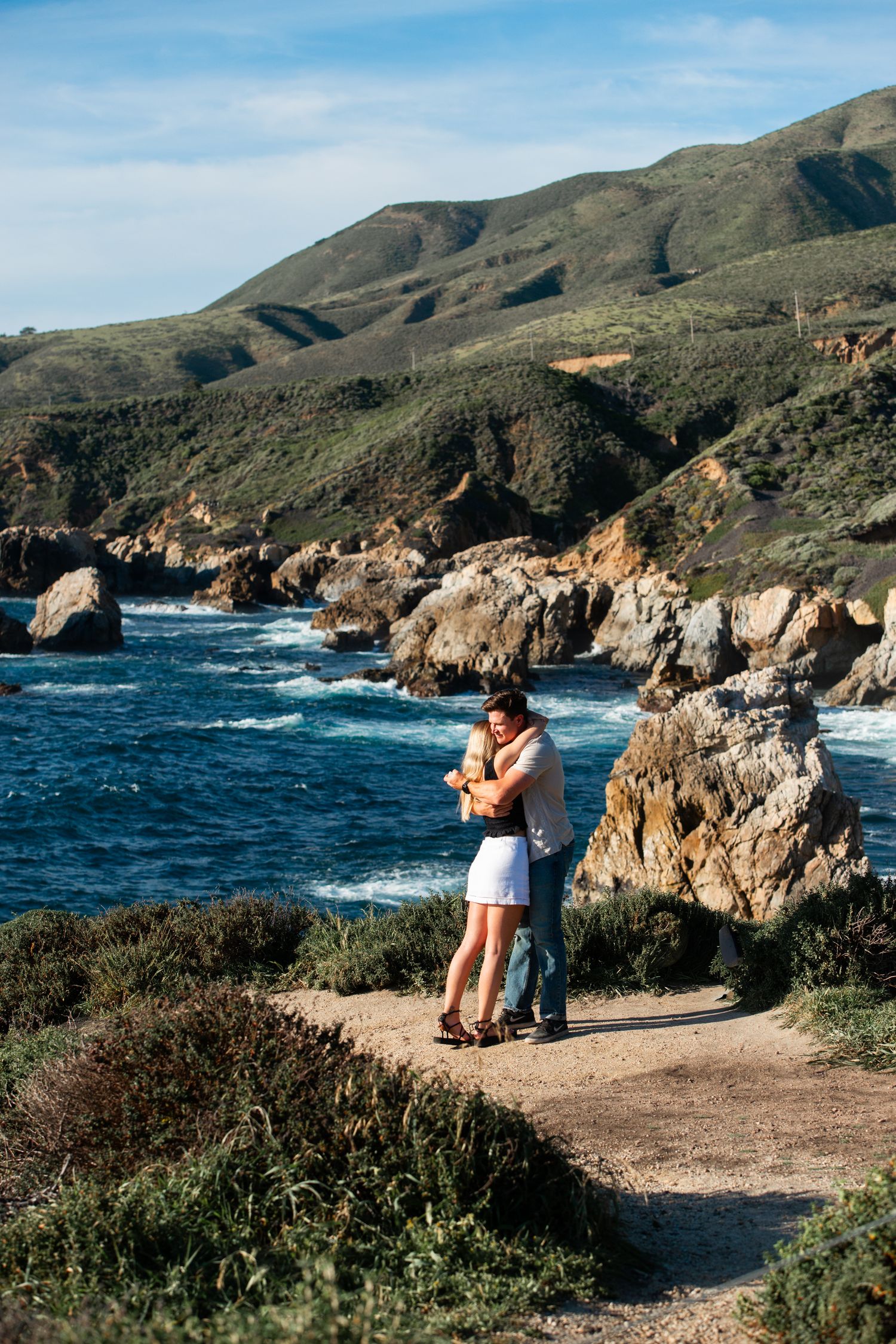 Couple embracing on a coastal path with rocky cliffs, blue ocean waves, and green hills in the background.