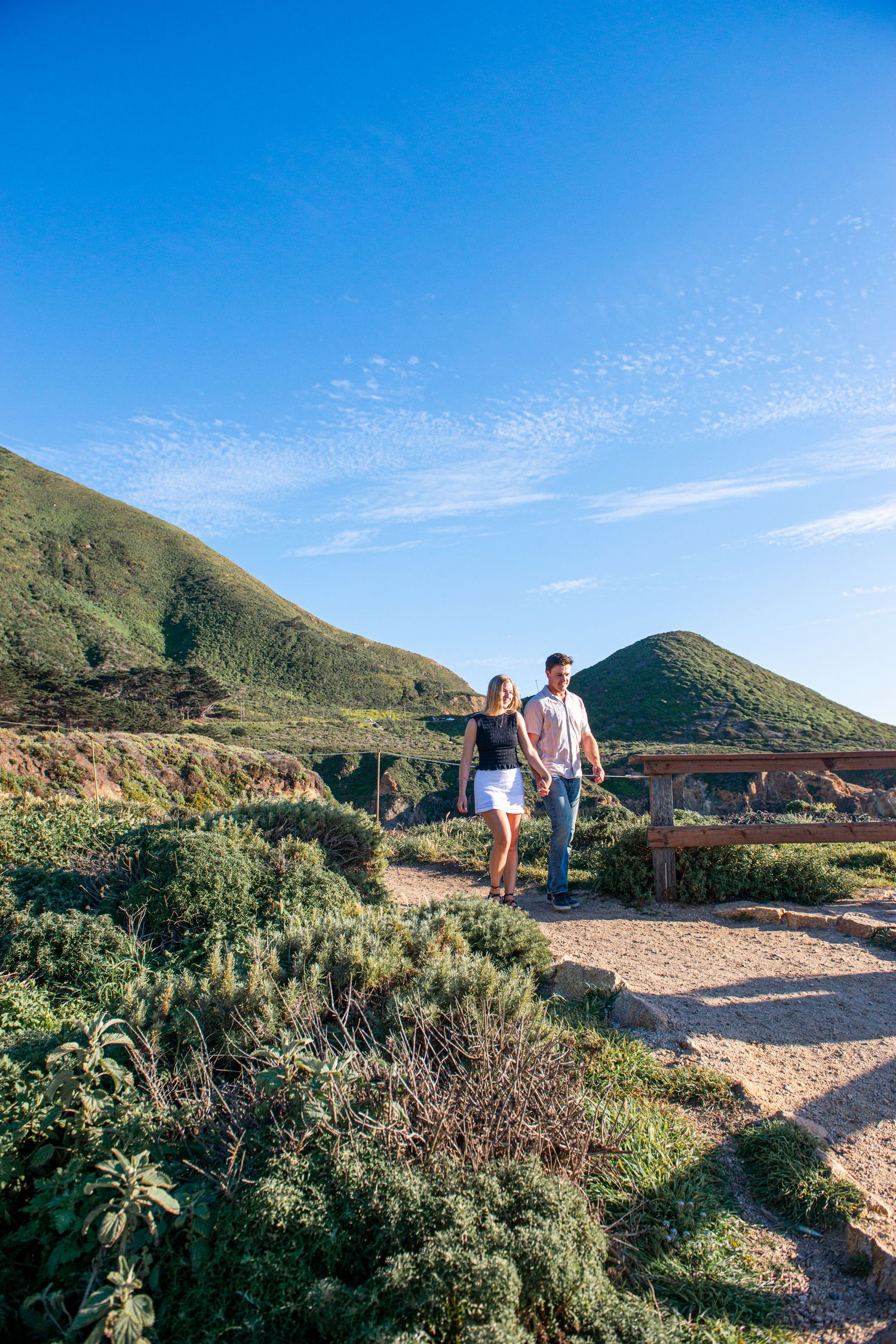 Couple holding hands walking on a scenic coastal trail with green hills and blue sky in Big Sur, California.