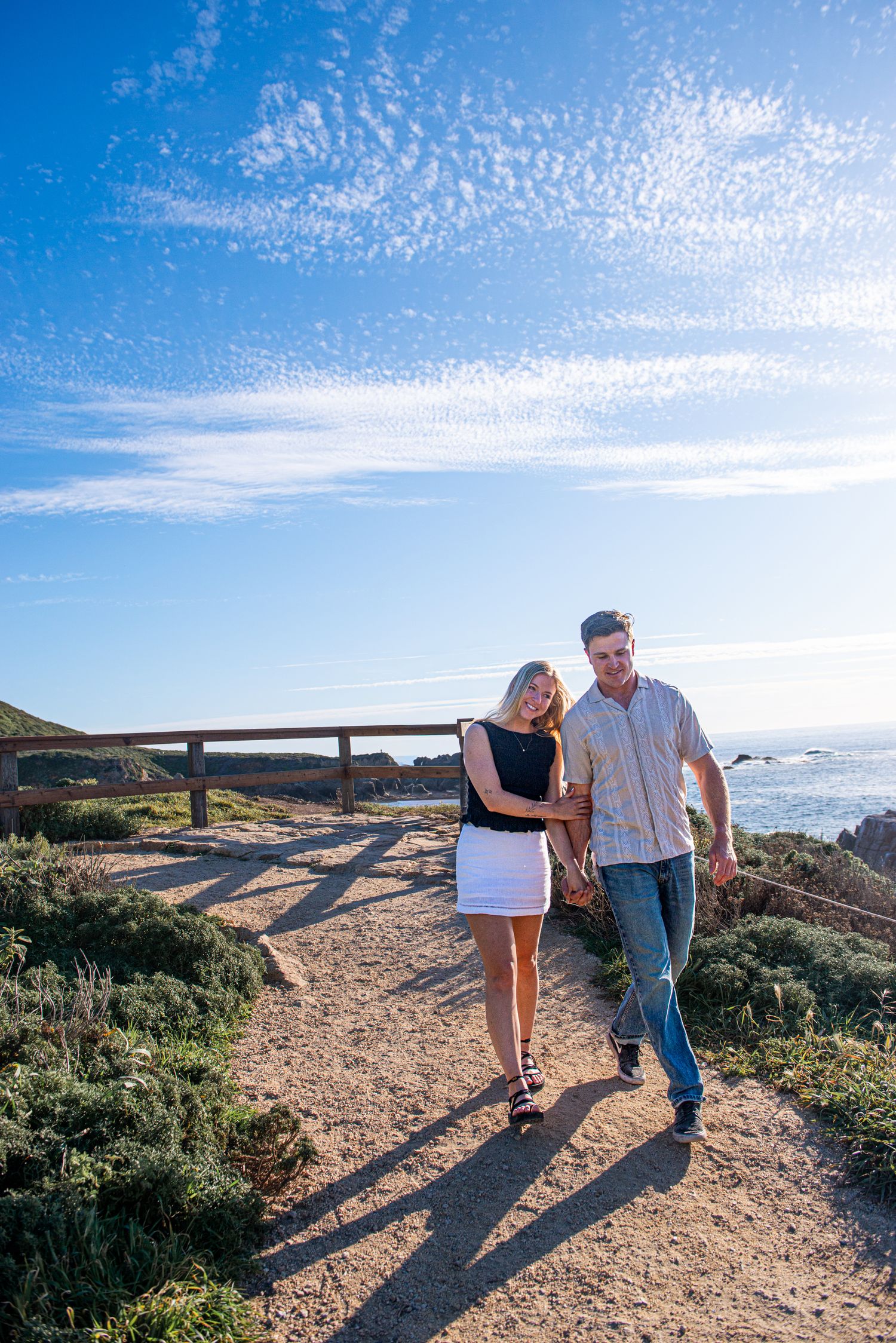Couple walking hand-in-hand on a coastal path with ocean views and blue sky.