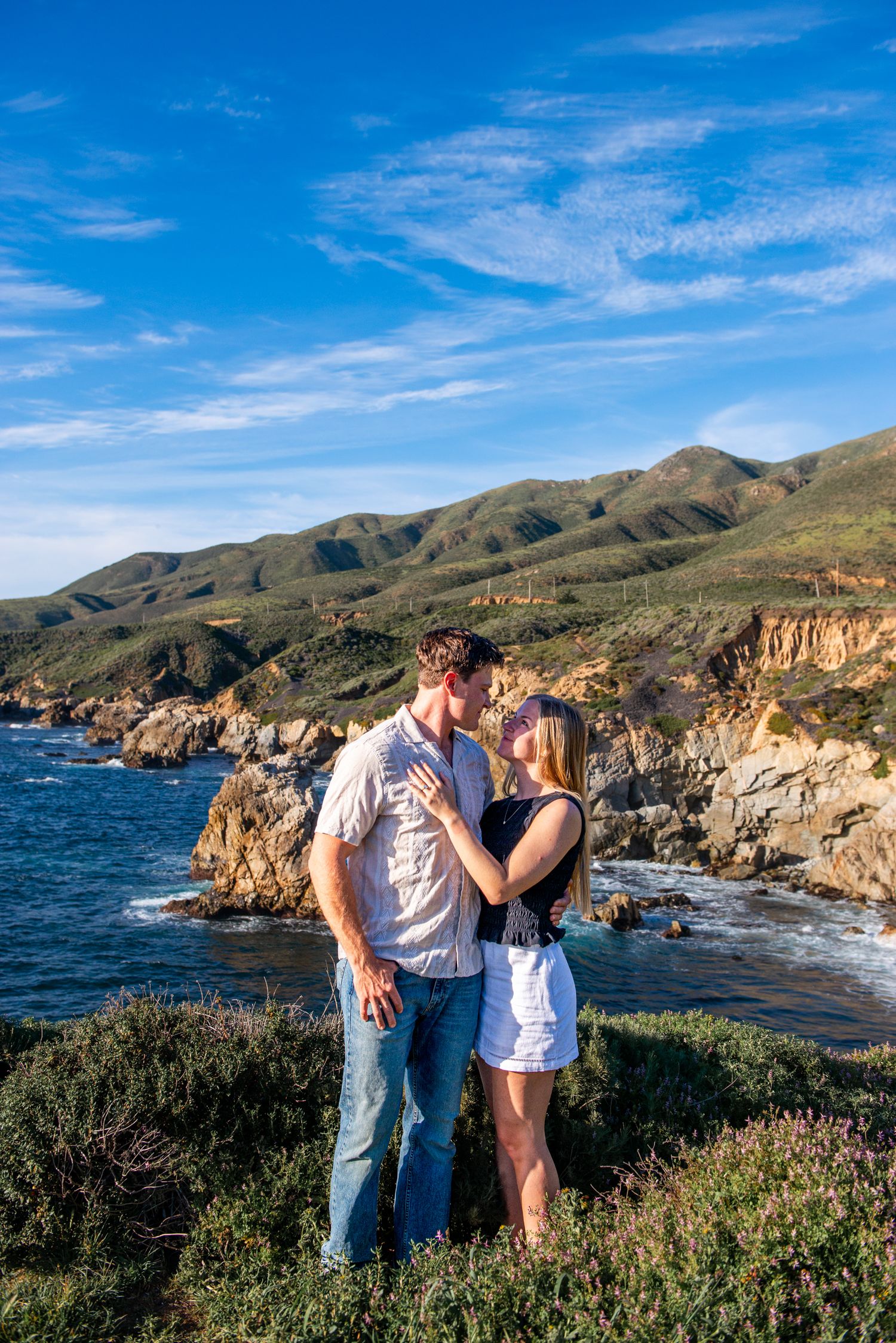 Romantic couple kissing on a scenic Big Sur coastline with mountains and ocean.