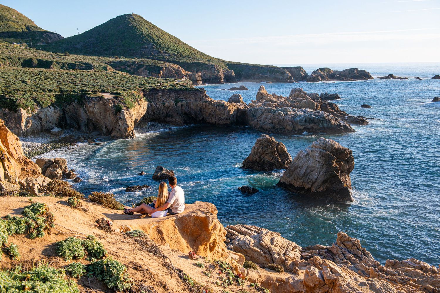Couple sitting on rocky coastal cliffs overlooking turquoise ocean waters with rugged sea stacks and green hills.