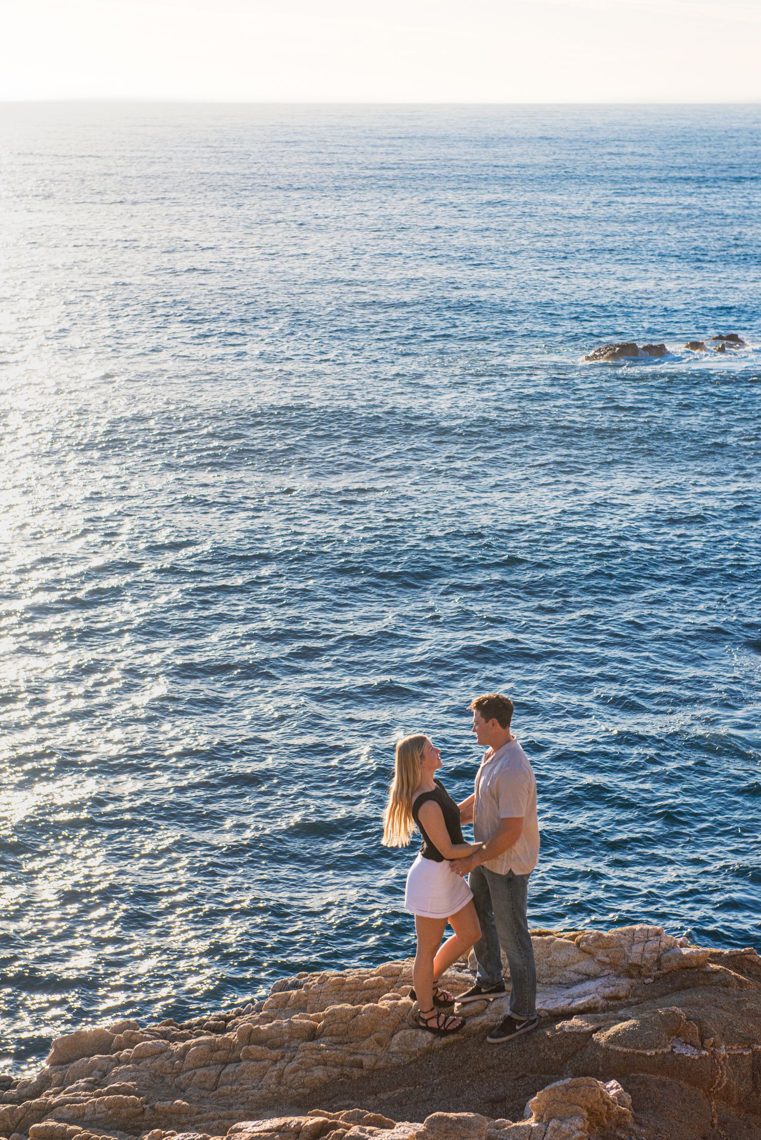 Couple standing on rocky coastal cliffs overlooking a shimmering blue ocean at sunset.
