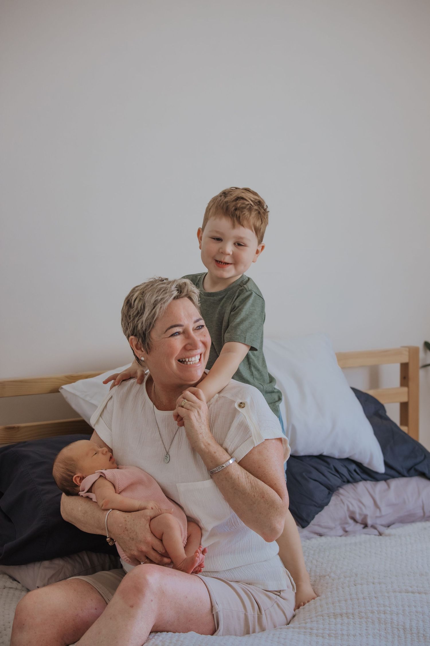 A smiling woman sits on a bed holding a newborn while a young boy in a green shirt hugs her from behind during in home newborn photoshoot.