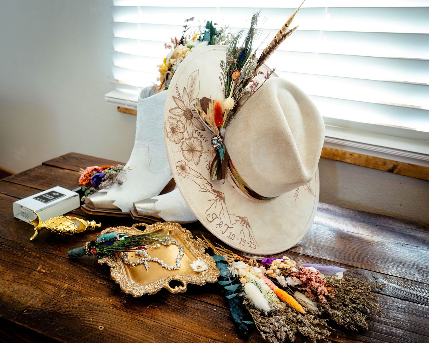 White cowboy hat and boots with floral embroidery displayed on wooden surface with vintage tray and dried flowers.