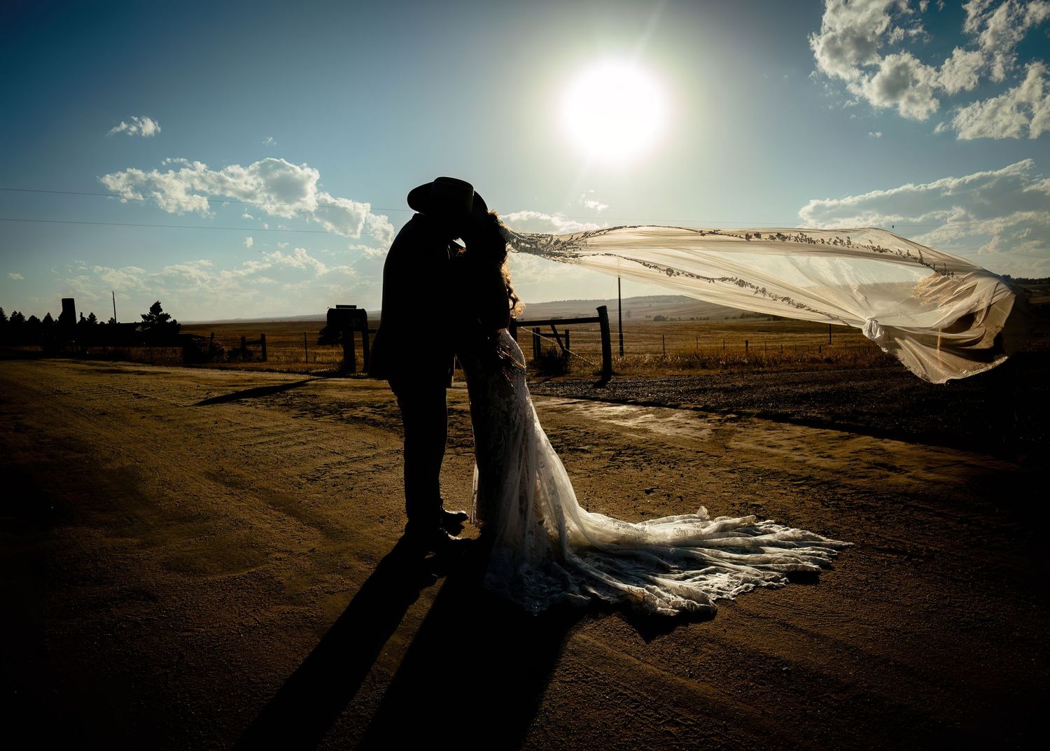 Silhouette of bride and groom kissing at sunset on a dirt road, with wedding veil flowing in the wind.