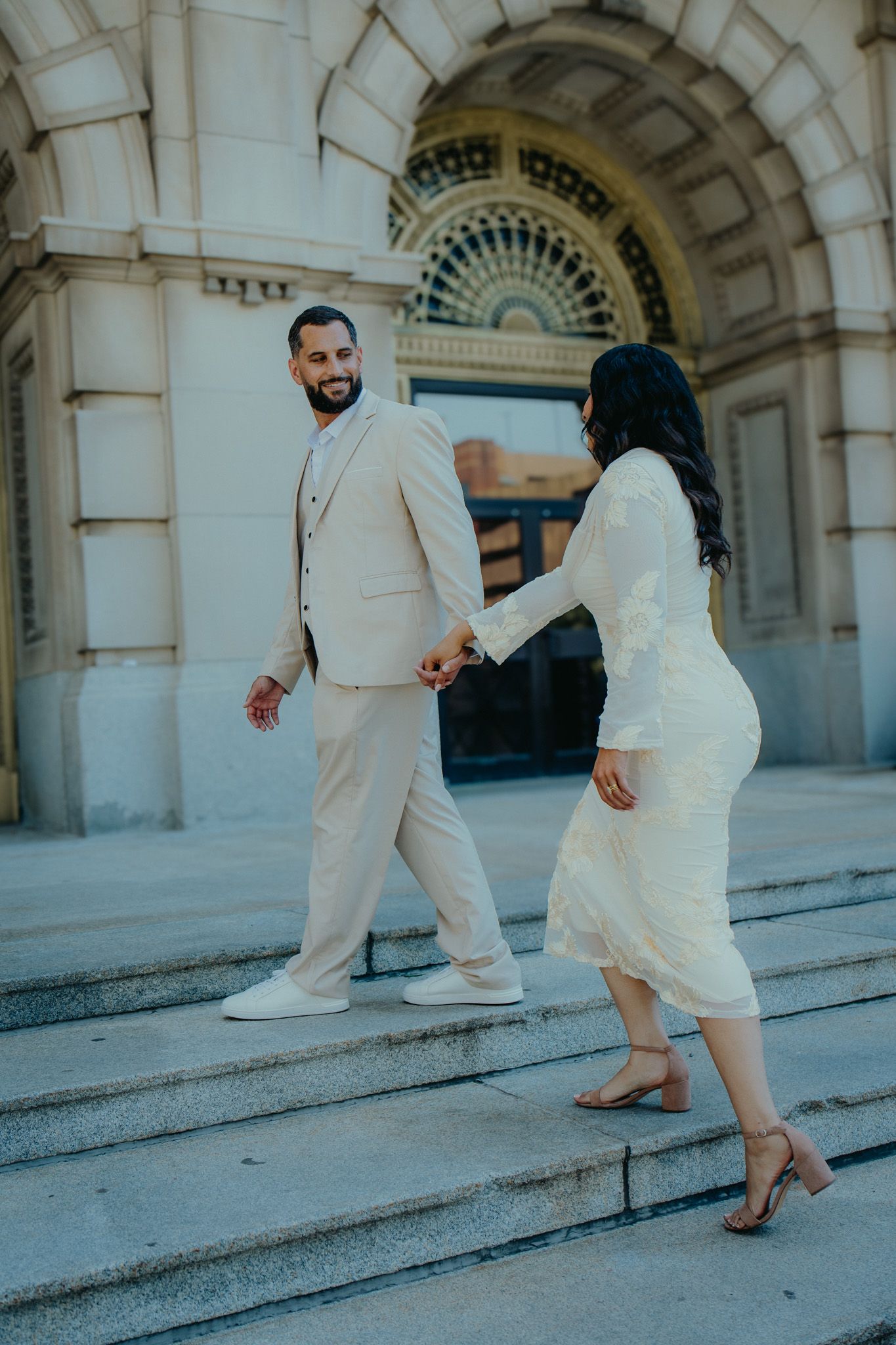 Couple in matching cream outfits holding hands on stone steps outside an ornate building entrance.