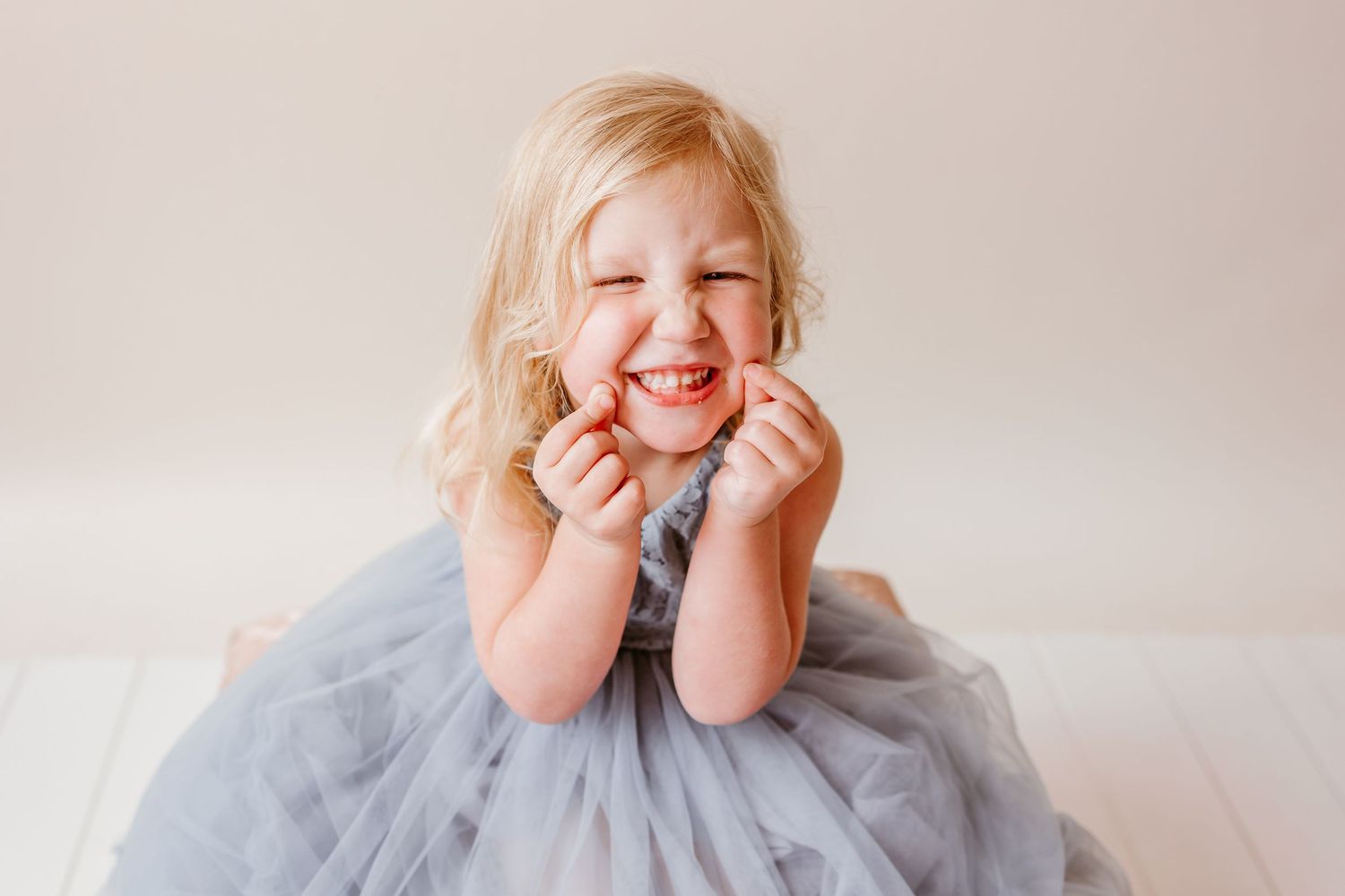 A joyful blonde toddler girl in a gray tulle dress laughs with hands on cheeks against a white background.
