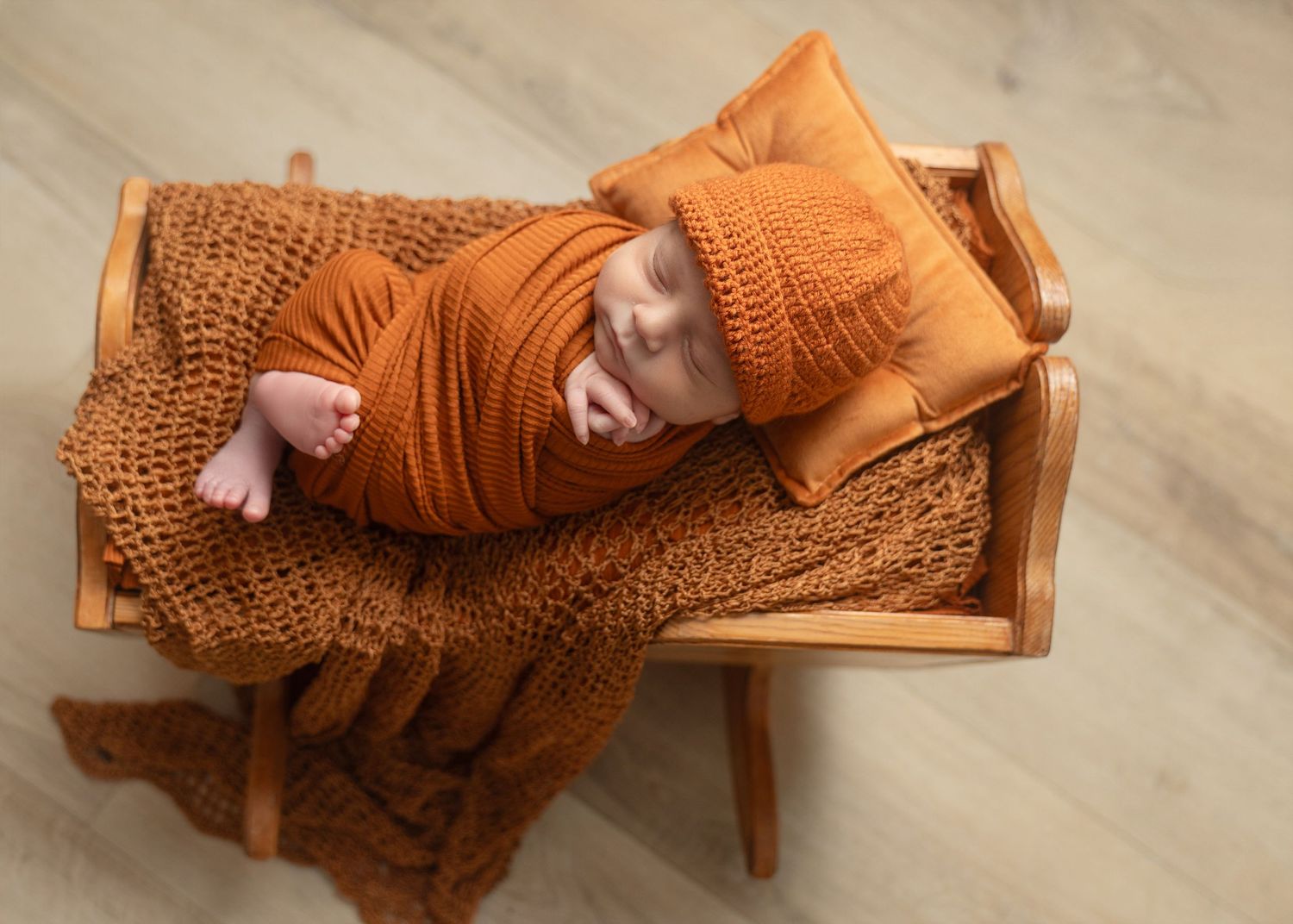 Newborn baby wrapped in orange knit blanket and hat, sleeping peacefully on a miniature wooden bench.