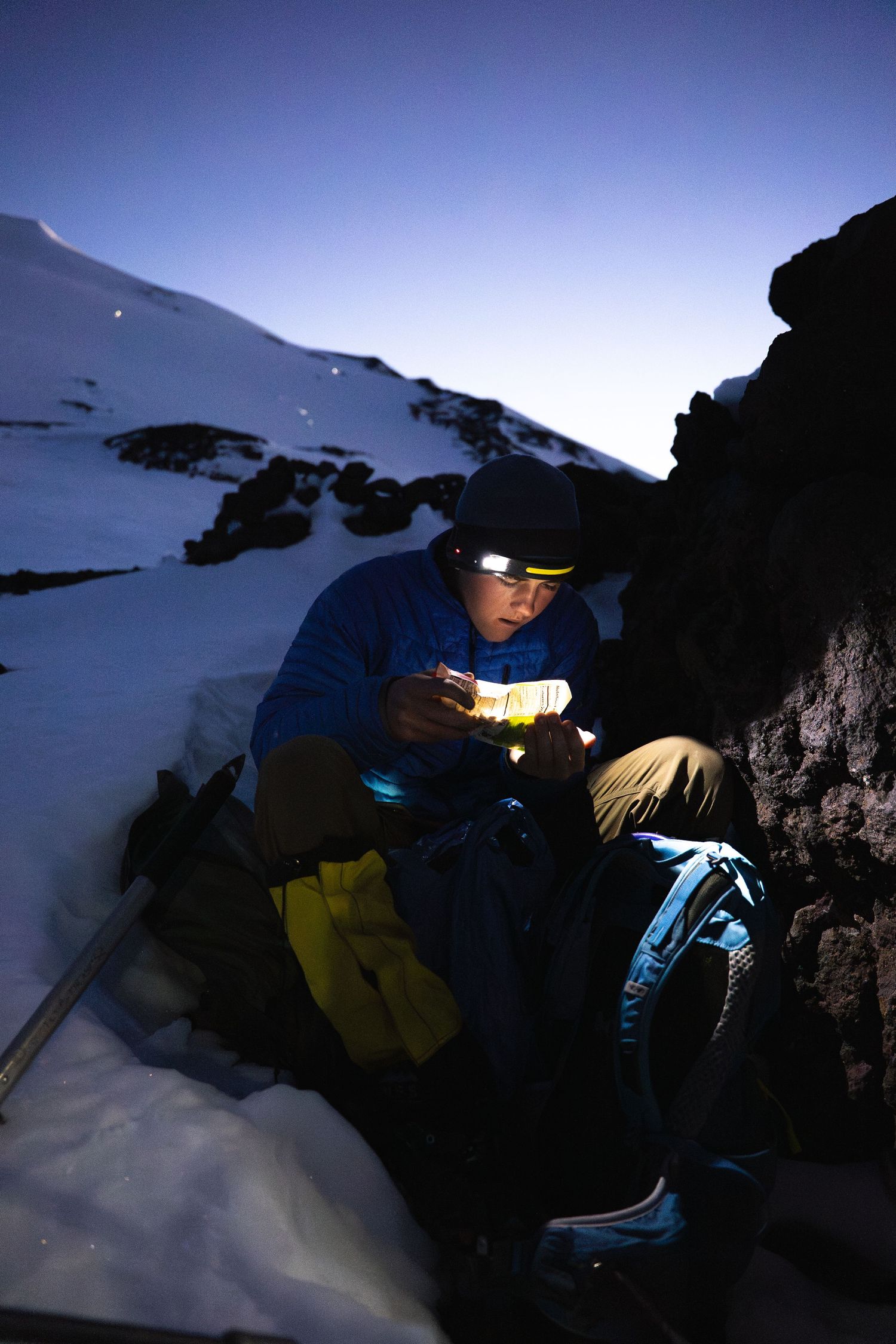 Mountain climber wearing headlamp sits against snowy rocks at dusk, reviewing gear during a winter alpine expedition.