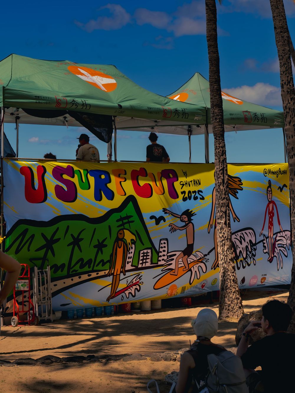 The U Surf Cup 2025 announcer's tent at Waikiki, draped with a bright hand-painted surf mural banner as spectators watch from the sand.