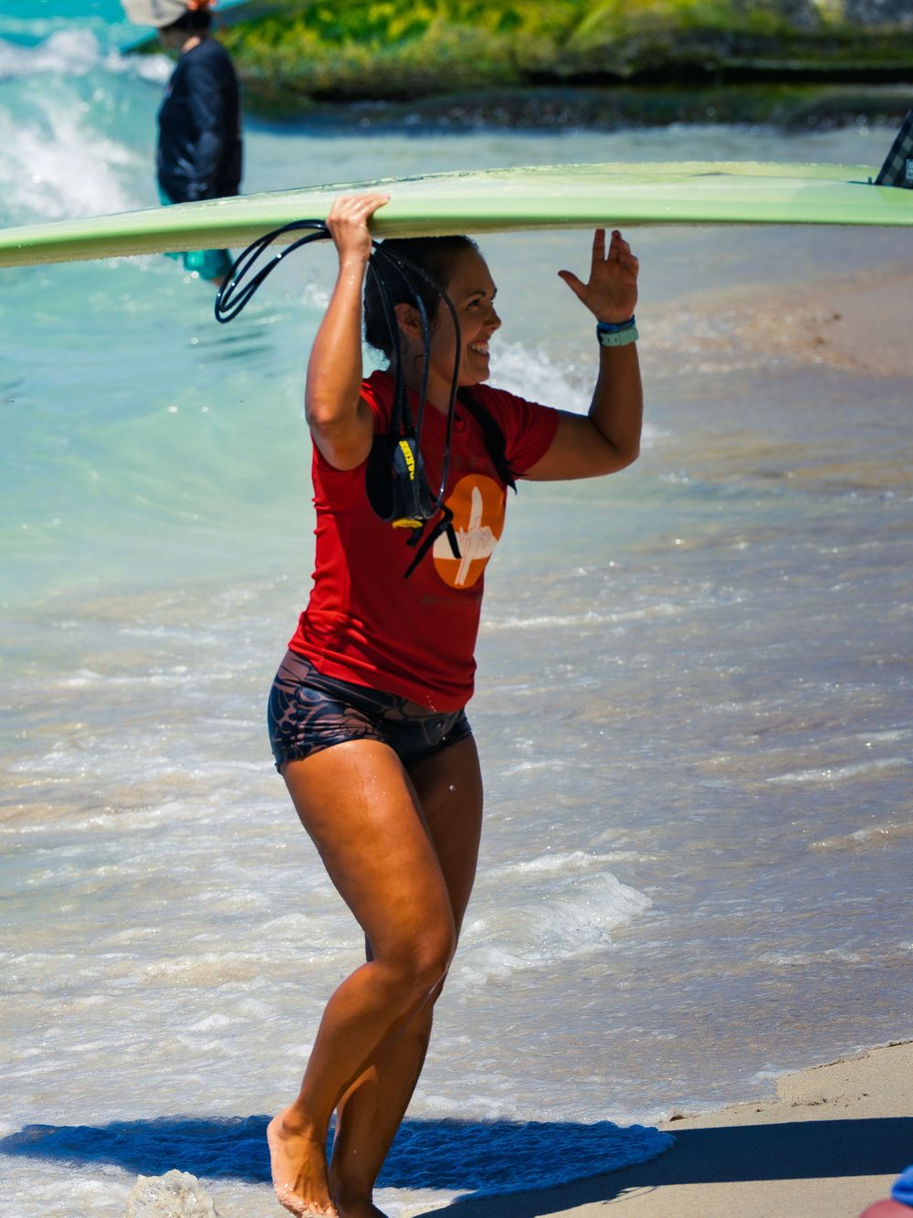 A smiling surfer in a red U Surf Cup shirt carries a green surfboard on her head out of the water at Waikiki, waving as she reaches the sand