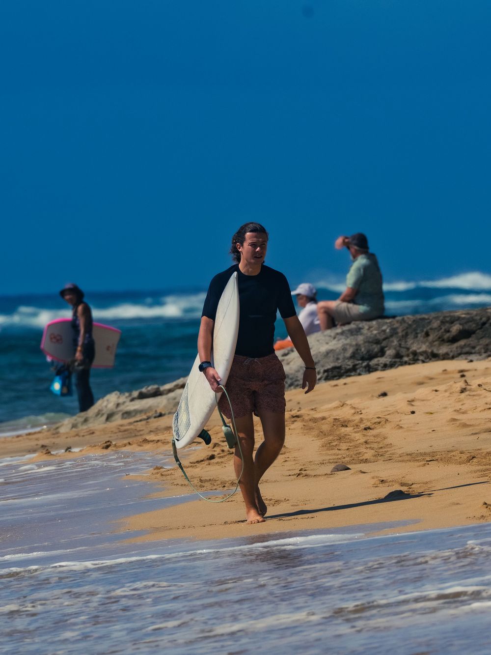 A surfer in a black top walks up the sand at Waikiki carrying a white surfboard, other beachgoers and the blue ocean behind him.