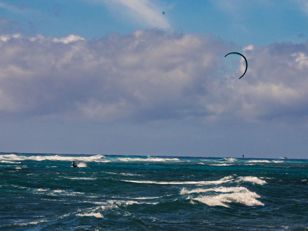 A kitesurfer skims across choppy blue-green water at Waikiki, a curved kite arcing high against a bright sky of scattered clouds.