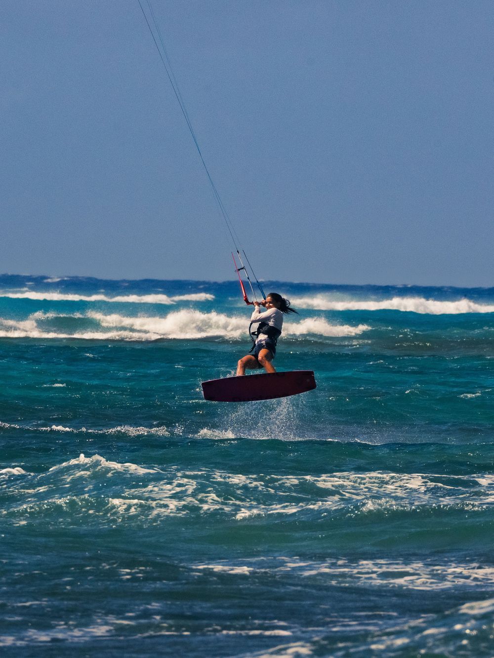 A kitesurfer catches air above the water at Waikiki, board lifting off a red deck as she grips the control bar, waves breaking behind.
