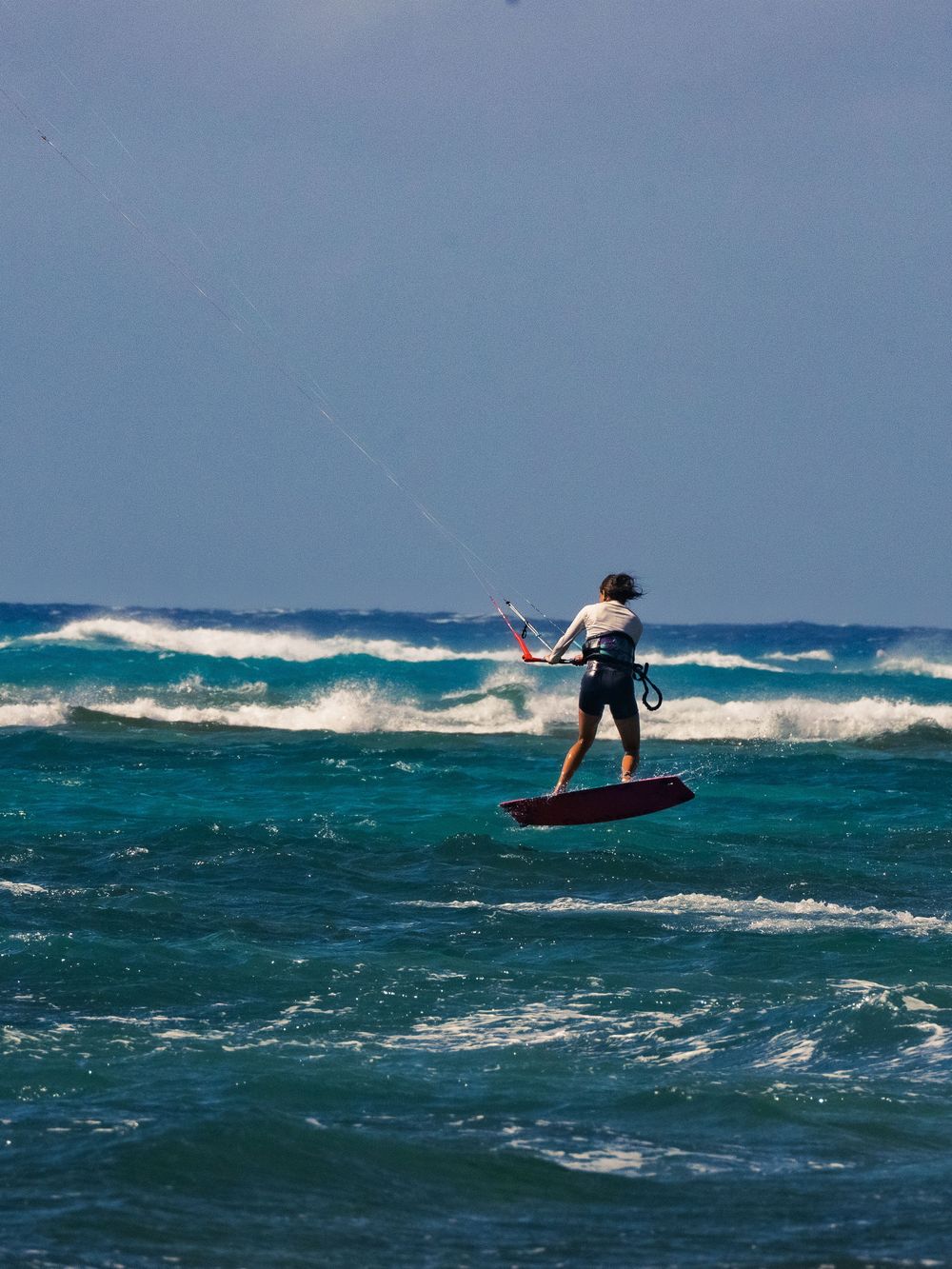 A kitesurfer seen from behind lifts a red board off the water at Waikiki, gripping the control bar as breaking waves roll across the horizon