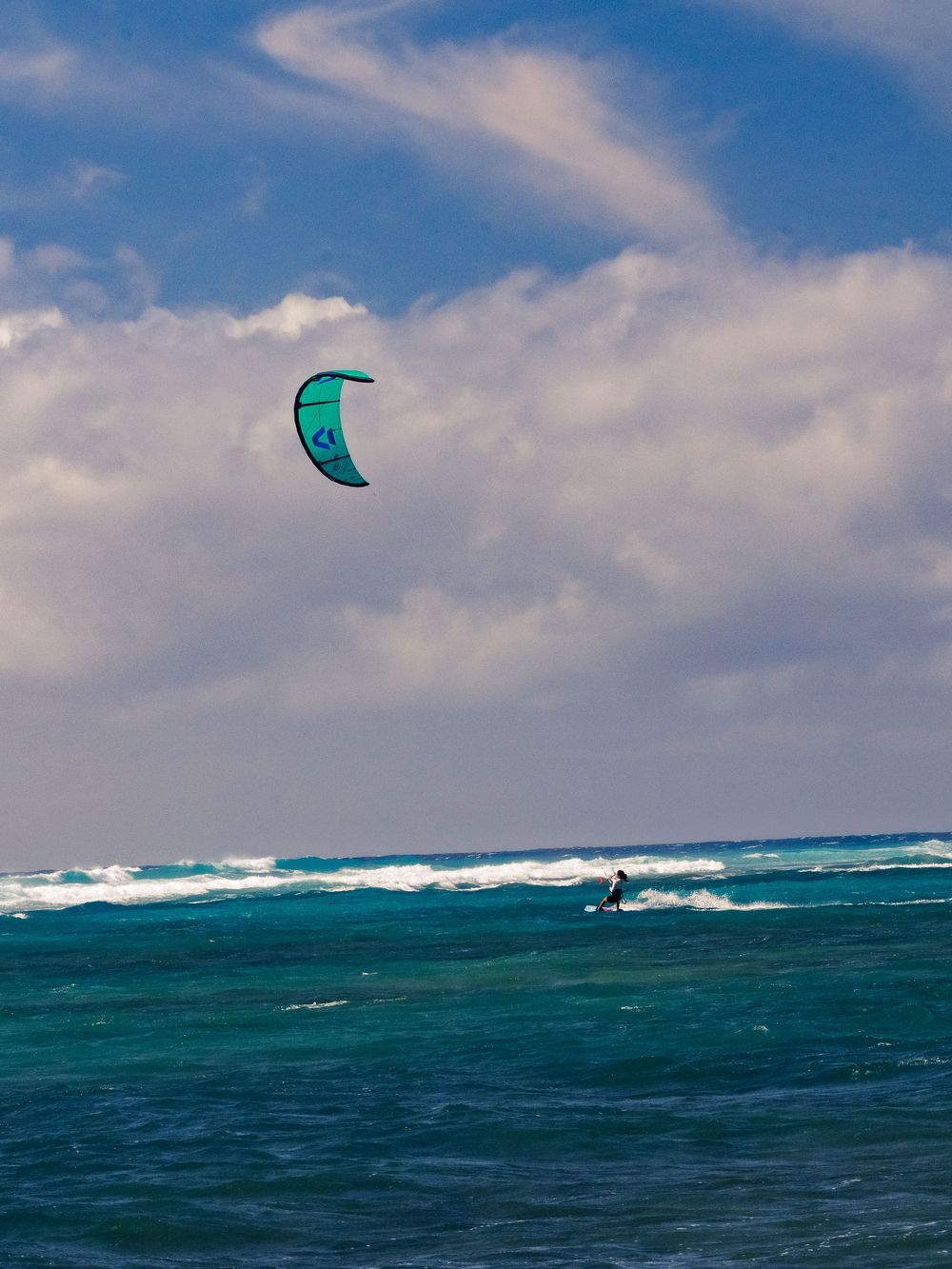 A kitesurfer rides across turquoise water at Waikiki, a teal kite lofted high on long lines against a blue sky streaked with white clouds.