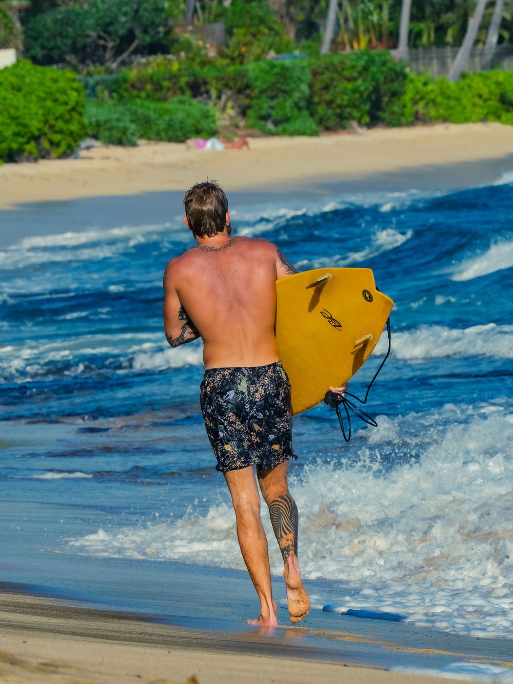 A shirtless bodyboarder in floral boardshorts runs into the shorebreak at Waikiki with a yellow bodyboard, waves breaking and sand behind.